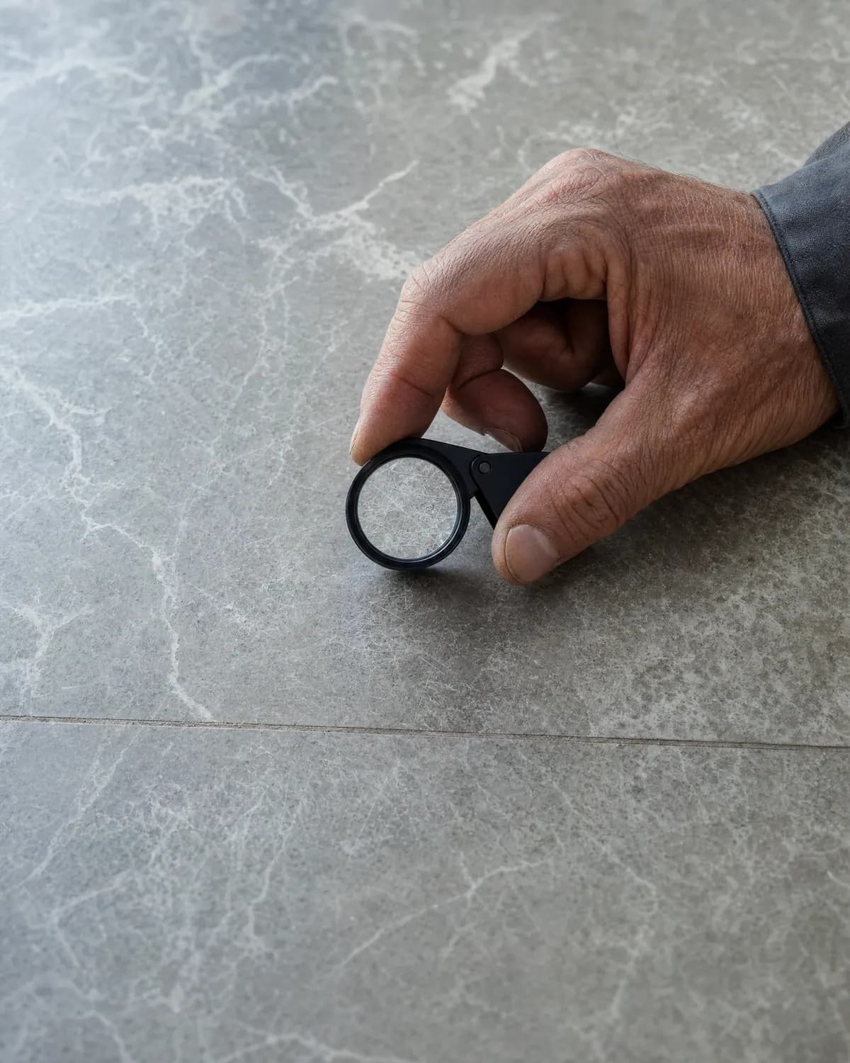 Hand hält eine kleine Lupe dicht über einen hellgrauen Marmorboden mit feinen Kratzern